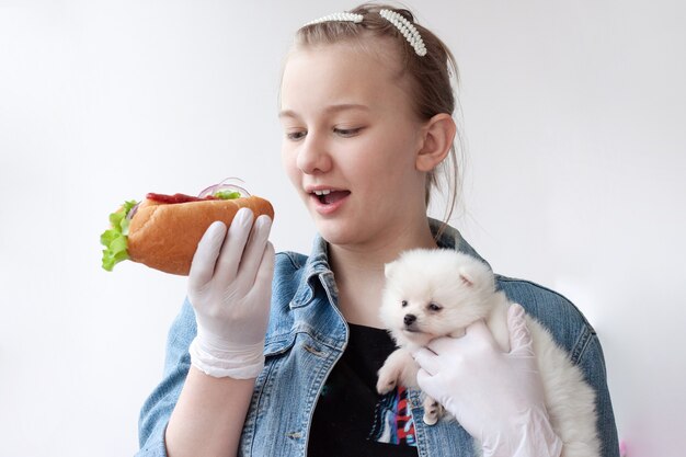 Ein junges Mädchen mit hellem Haar in einer blauen Jeansjacke und medizinischen Handschuhen hält in der einen Hand einen Hot Dog und in der anderen einen kleinen weißen Pommerschen Hund.