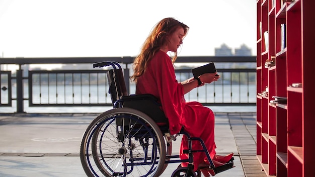 Ein junges Mädchen mit roten Haaren in einem roten Kleid sitzt im Rollstuhl und liest ein Buch