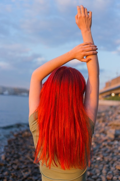 Ein junges Mädchen mit roten lockeren Haaren mit erhobenen Händen posiert in den Sonnenuntergangstrahlen für die Kamera