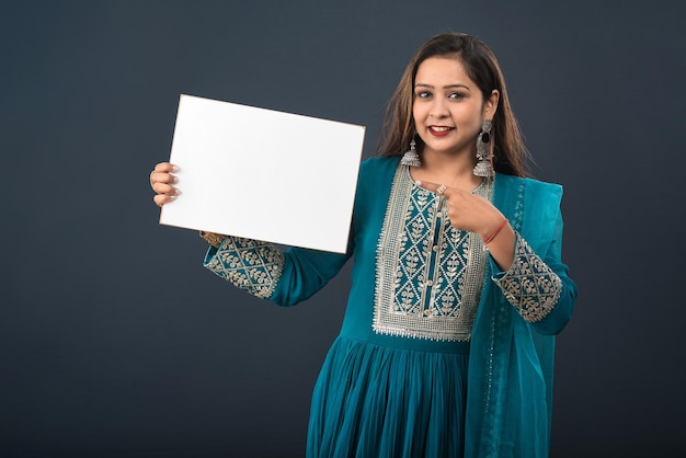 Ein junges Mädchen oder eine junge Frau, die ein traditionelles indisches Kleid trägt und ein Schild in ihren Händen auf grauem Hintergrund hält