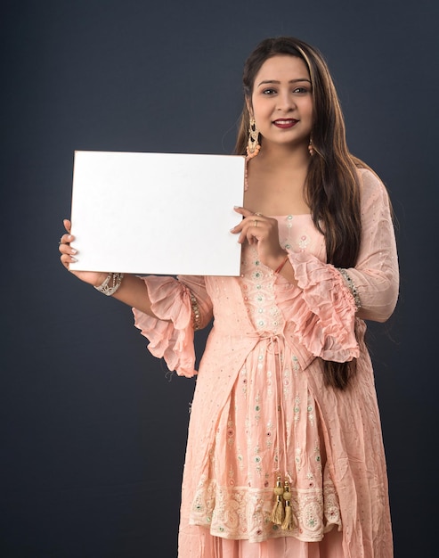 Ein junges Mädchen oder eine junge Frau, die ein traditionelles indisches Kleid trägt und ein Schild in ihren Händen auf grauem Hintergrund hält