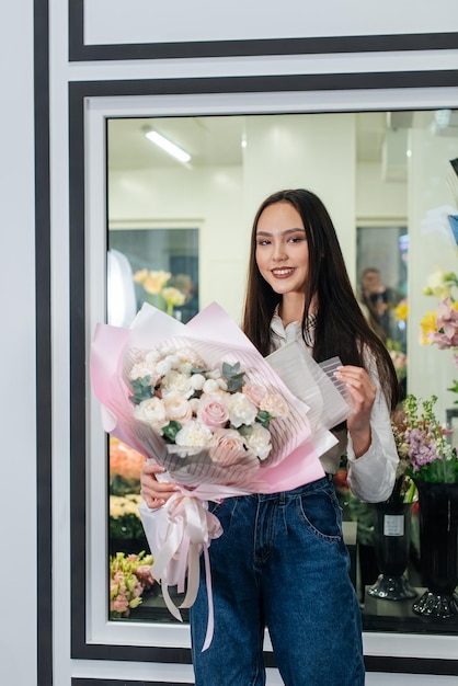 Ein junges mädchen posiert mit einem schönen festlichen blumenstrauß vor dem hintergrund eines gemütlichen blumenladens floristik und blumenstraußherstellung in einem blumenladen kleines unternehmen