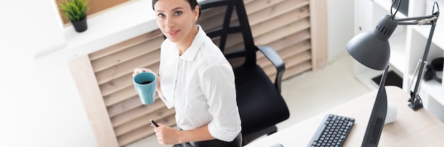 Ein junges Mädchen setzte sich auf einen Tisch im Büro, hielt eine Tasse in der Hand und arbeitete