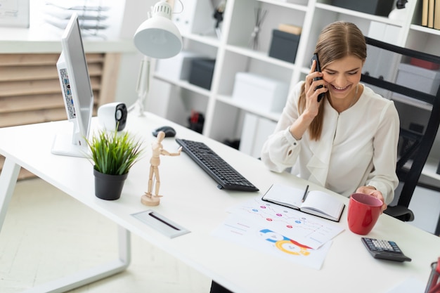 Ein junges Mädchen sitzt an einem Tisch im Büro, hält eine rote Tasse in der Hand und telefoniert.