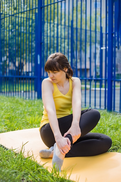 Ein junges Mädchen sitzt auf einem Teppich und zuckt vor Schmerz zusammen. Das Mädchen sitzt auf einer gelben Matte im Gras und reibt sich das schmerzende Bein.