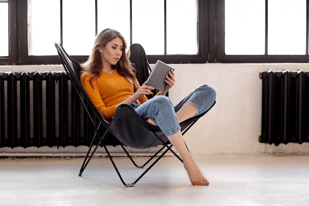 Ein junges Mädchen sitzt zu Hause in einem eleganten schwarzen Ledersessel, hält eine Tasse Kaffee und eine Tablette in der Hand. Arbeit und Entspannung zu Hause