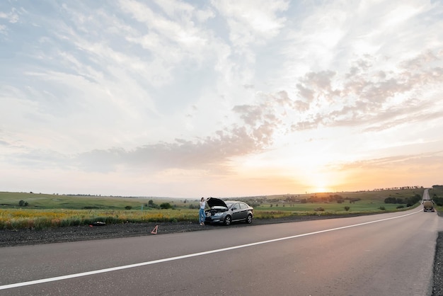 Ein junges Mädchen steht bei Sonnenuntergang in der Nähe eines kaputten Autos mitten auf der Autobahn und versucht, am Telefon um Hilfe zu rufen Warten auf Hilfe Autoservice Autopanne auf der Straße