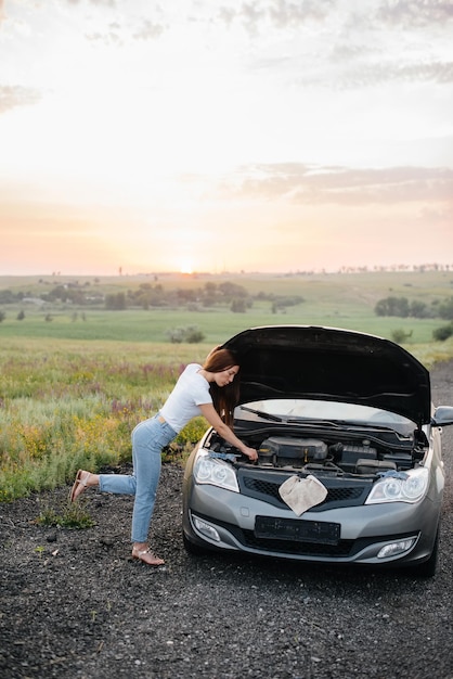 Ein junges Mädchen steht bei Sonnenuntergang in der Nähe eines kaputten Autos mitten auf der Autobahn und versucht, es zu reparieren. Panne und Reparatur des Autos. Beheben des Problems.