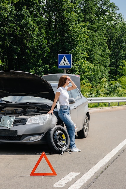 Ein junges Mädchen steht in der Nähe eines kaputten Autos mit einem kaputten Rad mitten auf der Autobahn und wartet frustriert an einem heißen Tag auf Hilfe. Panne und Panne des Autos. Warten auf Hilfe.