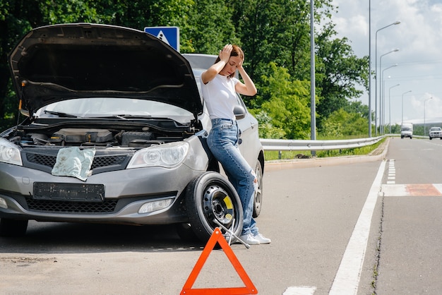 Ein junges Mädchen steht in der Nähe eines kaputten Autos mit einem kaputten Rad mitten auf der Autobahn und wartet frustriert an einem heißen Tag auf Hilfe. Panne und Panne des Autos. Warten auf Hilfe.