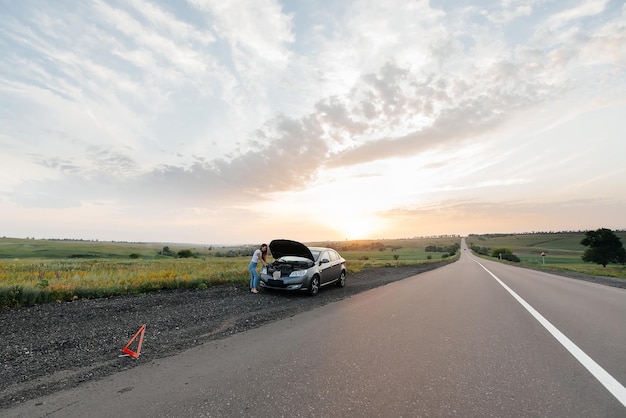 Ein junges Mädchen steht während des Sonnenuntergangs in der Nähe eines kaputten Autos mitten auf der Autobahn und versucht, es zu reparieren Fehlerbehebung des Problems Warten auf Hilfe Autoservice Autopanne auf der Straße