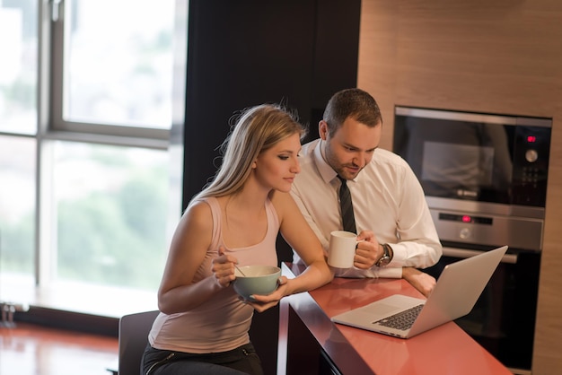 Ein junges Paar bereitet sich auf den Job vor und benutzt einen Laptop. Der Mann trinkt Kaffee, während die Frau gemeinsam in einem Luxushaus frühstückt, lächelnd auf den Bildschirm schaut.