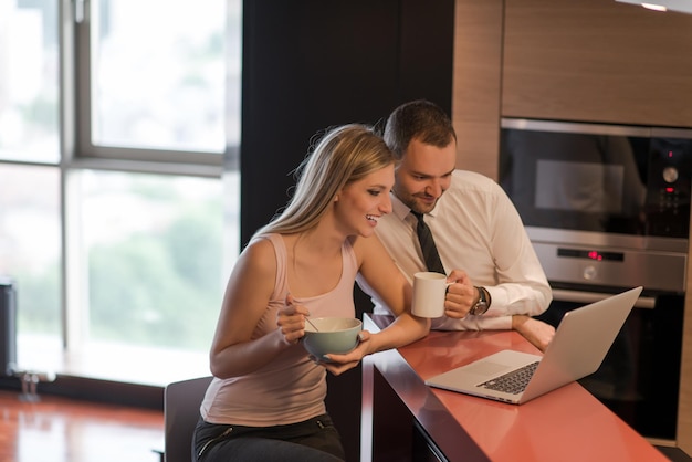 Ein junges Paar bereitet sich auf den Job vor und benutzt einen Laptop. Der Mann trinkt Kaffee, während die Frau gemeinsam in einem Luxushaus frühstückt, lächelnd auf den Bildschirm schaut.