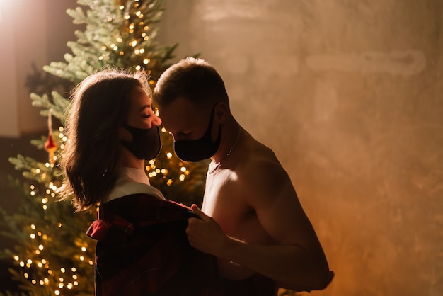 Ein junges Paar Mann und Frau in medizinischen Einwegmasken unter einem Weihnachtsbaum.