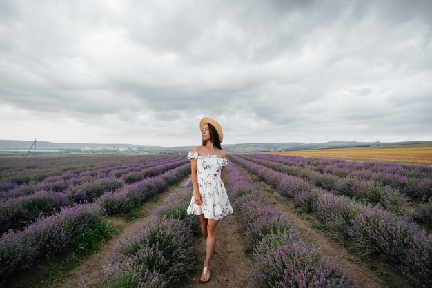 Ein junges schönes Mädchen in einem zarten Kleid und Hut geht durch ein wunderschönes Lavendelfeld und genießt den Duft von Blumen. Urlaub und schöne Natur.