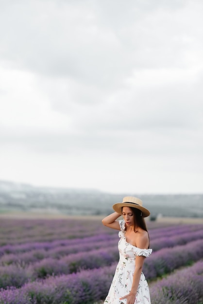 Ein junges schönes Mädchen in einem zarten Kleid und Hut geht durch ein wunderschönes Lavendelfeld und genießt den Duft von Blumen. Urlaub und schöne Natur.