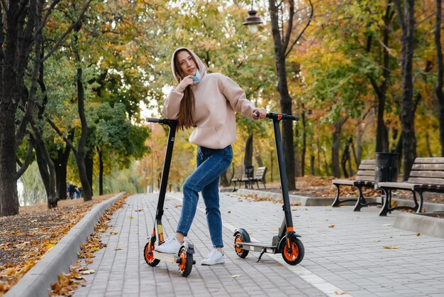 Ein junges schönes Mädchen in einer Maske fährt an einem warmen Herbsttag auf einem Elektroroller im Park. Im Park spazieren gehen.