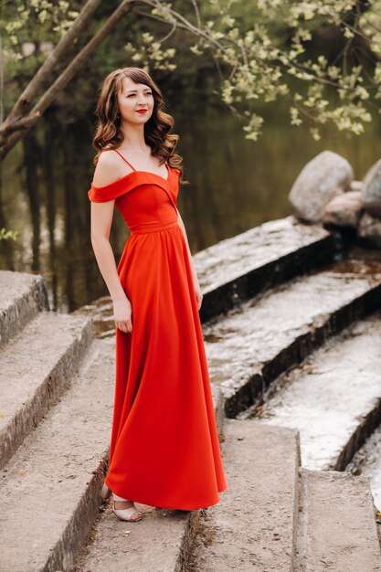 Ein junges schönes Mädchen mit langen braunen Haaren in einem langen roten Kleid kommt die Treppe in der Nähe des Sees herunter.