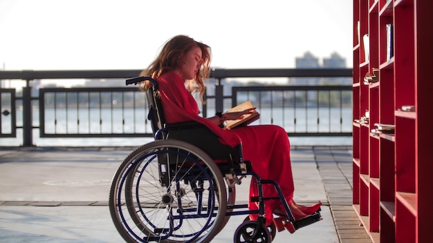 Ein junges schönes Mädchen mit roten Haaren in einem roten Kleid sitzt im Rollstuhl und liest ein Buch