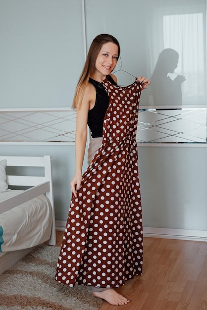 Ein junges schönes Mädchen wählt ein Outfit im Kleiderschrank zu Hause im Schlafzimmer. Eine Fashionista hält einen Kleiderbügel mit einem langen Kleid mit gepunktetem Lebensstil