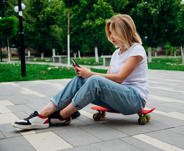 Ein junges schönes Teenager-Mädchen sitzt an einem schönen Sommertag im Park auf einem Skateboard. Ein Mädchen spricht per Video auf einem Handy im Freien. Chatten Sie mit Freunden