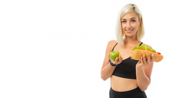 Ein junges Sportmädchen mit blonden Haaren in einem schwarzen Sportoberteil und schwarzen Leggings hält einen grünen Apfel und ein Sandwich.