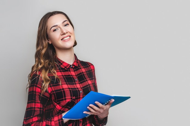 Ein junges Studentenmädchen in einem karierten Hemd hält ein Notizbuch an einer grauen Wand mit für Text. Netter Student mit Dokumentenpapieren in den Händen. Mädchen lernt Englisch.