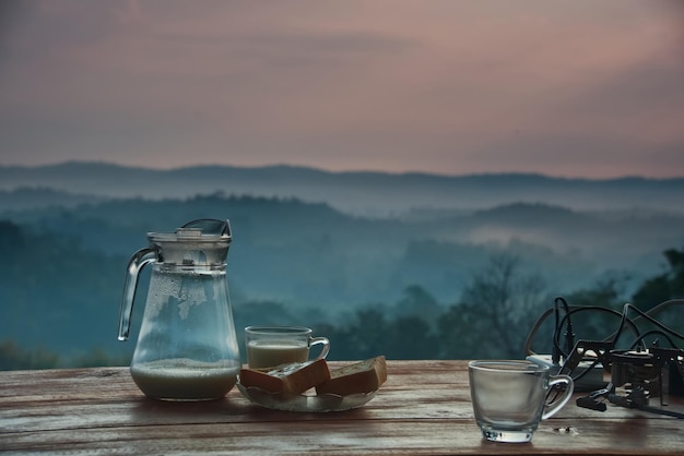 Ein Kaffeemilchbecher und ein Brot auf einem Holztisch mit verschwommenem Bergwaldhintergrund an einem Morgen