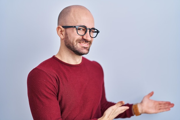 Ein kahlköpfiger junger Mann mit Bart, der über einem weißen Hintergrund steht, eine Brille trägt, die zum Betreten einlädt, lächelt natürlich mit offener Hand