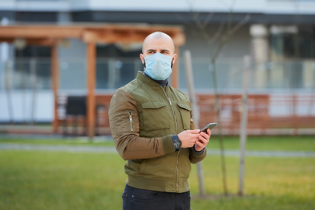 Ein kahlköpfiger Mann in einer medizinischen Gesichtsmaske, um zu vermeiden, dass das verbreitete Coronavirus sein Smartphone in der gemütlichen Straße überprüft.