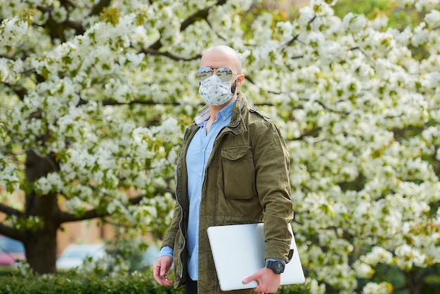 Ein kahlköpfiger Mann mit Bart in einer medizinischen Gesichtsmaske, um die Ausbreitung des Coronavirus zu vermeiden, geht mit einem Laptop im Park spazieren. Ein Mann trägt eine n95 Gesichtsmaske und eine Pilotensonnenbrille im Garten zwischen blühenden Bäumen.