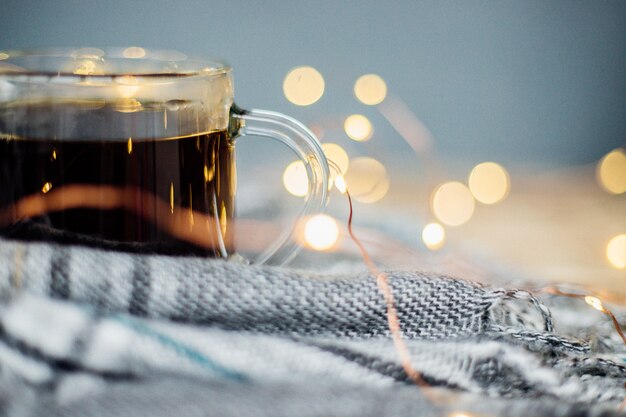 Ein karierter Schal mit Fransen liegt auf hellem Holzhintergrund Ein Glasbecher mit schwarzem Kaffee