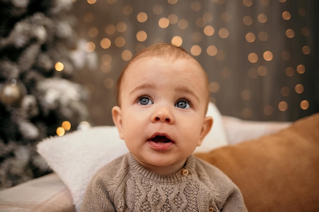 Ein kaukasischer Junge mit blauen Augen in einem beigen Strickpullover sitzt auf dem Sofa auf einem Neujahrshintergrund