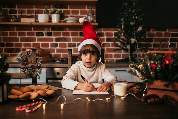 Ein kaukasischer kleiner Junge in einem weißen Strickpullover und einer roten Neujahrsmütze sitzt auf dem Küchentisch und schreibt eine Neujahrsbotschaft