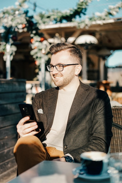 Foto ein kaukasischer mann mit brille hält smartphone und trinkt kaffee aus einer tasse im café. fernarbeit per smartphone. stadt leben. geschäftsperson in der pause. beiläufige art und weise. erfolgskonzept. gemischte medien.