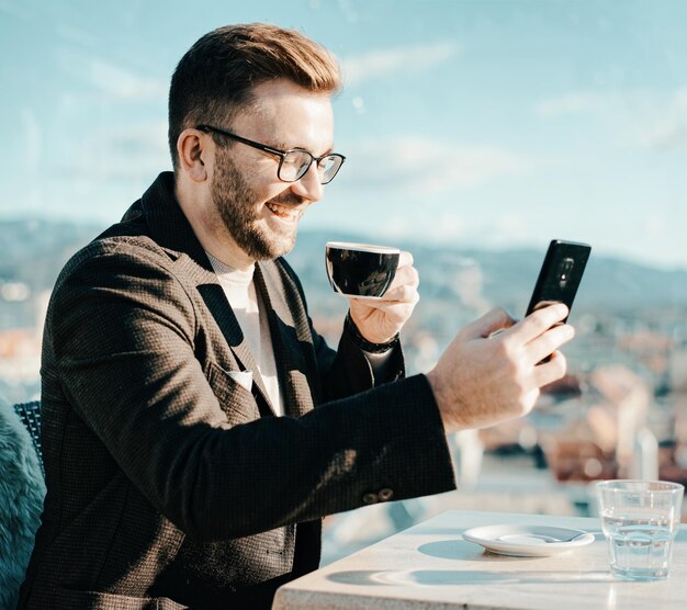 Foto ein kaukasischer mann mit brille hält smartphone und trinkt kaffee aus einer tasse im café. fernarbeit per smartphone. stadt leben. geschäftsperson in der pause. beiläufige art und weise. erfolgskonzept. gemischte medien.
