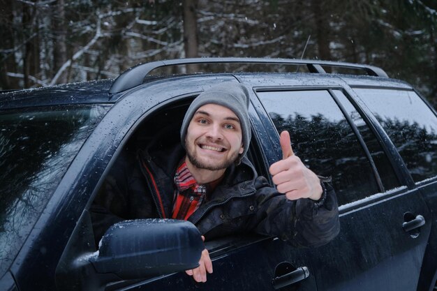 Ein kaukasischer Mann sitzt an einem verschneiten Wintertag am Steuer seines Autos und zeigt die Geste „Daumen hoch“.