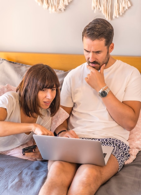 Ein kaukasisches Paar auf dem Bett mit einem Computer und einem Telefon, das eine Reservierung in einem Hotel oder Flug vornimmt, Urlaub organisiert, neue Technologien in der Familie. Suche nach den besten Angeboten