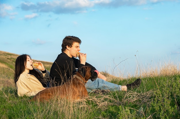 ein kerl und ein hübsches mädchen sitzen im gras und essen hamburger, ein hund ist bei ihnen