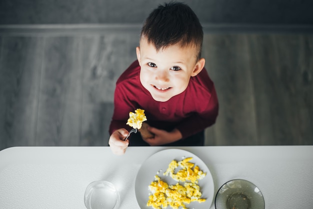 Ein Kind am Nachmittag in einer Weißlichtküche in einem burgunderfarbenen Pullover isst ein Omelett