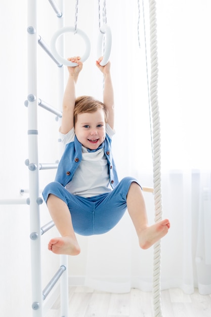 Ein Kind an der schwedischen Wand treibt zu Hause Sport, ein Junge klettert mit einem Seil auf eine Leiter, das Konzept von Sport und Gesundheit