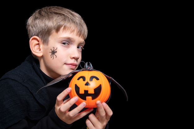 Ein Kind, das einen orangefarbenen kürbisförmigen Korb mit einem grinsenden Gesicht, Jacks Laterne und einer Fledermaus hält. Ein Junge mit einer Spinne auf der Wange wartet auf Halloween-Süßigkeiten. Happy Halloween-Konzept