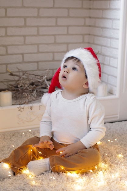 Ein Kind, das einen Weihnachtsmannhut trägt, sitzt an einem weißen Kamin mit Lichtern einer Weihnachtsgirlande und schaut auf.