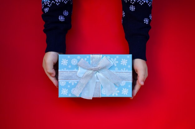 Ein Kind in einem Neujahrspullover hält eine Geschenkbox auf rotem Hintergrund Weihnachtsgeschenk