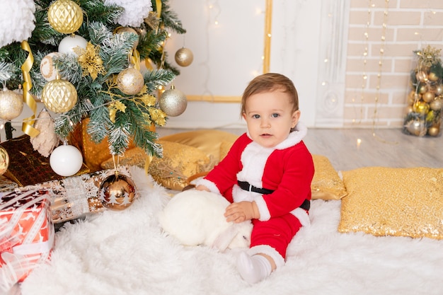 Ein Kind in einem Weihnachtsmannkostüm sitzt am Weihnachtsbaum mit einem Kaninchen, dem Konzept von Neujahr und Weihnachten