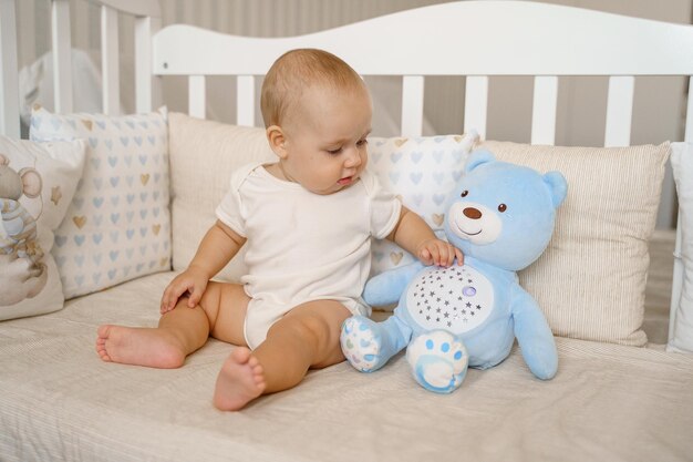 Ein Kind in einem weißen Body in einem weißen Kinderbett sitzt auf seinem Arsch neben einem Teddybär-Spielzeug