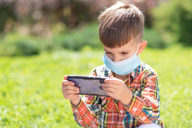 Ein Kind in einer medizinischen Maske sitzt im Gras und schaut ins Telefon