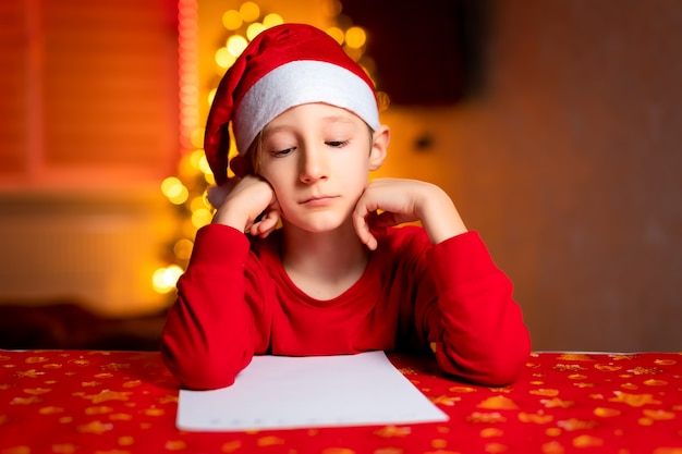 Ein Kind in einer roten Jacke auf dem Hintergrund eines Weihnachtsbaums und Girlanden schreibt einen Brief an den Weihnachtsmann