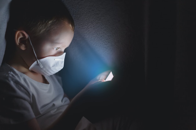 Ein Kind in einer Schutzmaske unter einer Decke spielt mit einem Smartphone und sitzt im Internet. Das Konzept, Zeit in sicherer Isolation zu verbringen.