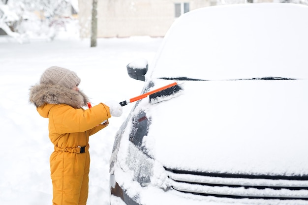 Ein Kind in Strickmütze und Handschuhen putzt vor der Fahrt Schnee vom Auto